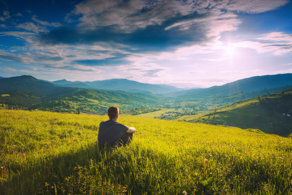 Tourist sitting on a hill in a fresh green grass and enjoy majestic sunset above carpathian mountain village.
