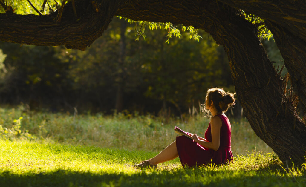 sunny portrait of a beautiful girl sitting on green glade under an arch of tree branches with book, woman reading novel on nature, concept hobby and lifestyle